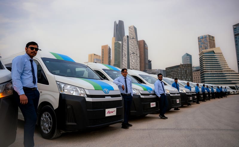 Uniformed Flow Mobility drivers beside a row of Toyota Hiace buses in front of the King Abdullah Financial District towers